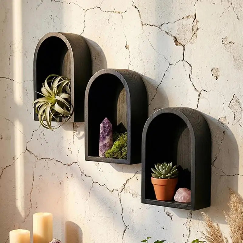 Three dark charcoal arched floating wall shelves mounted on a textured beige wall with cracks. The shelves are styled with decorative items including an air plant, a purple crystal, moss, a small potted succulent, and a pink stone.