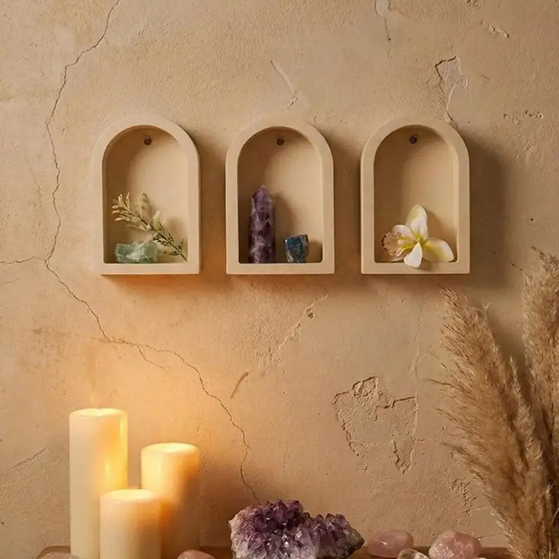 Three arched wooden wall niches mounted on a textured beige plaster wall, styled with crystals, a dried flower, and lit candles below.