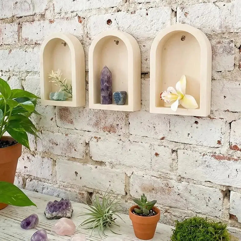 Three light beige arched wall niches mounted on a white brick wall, styled with crystals, a small orchid, and potted plants.