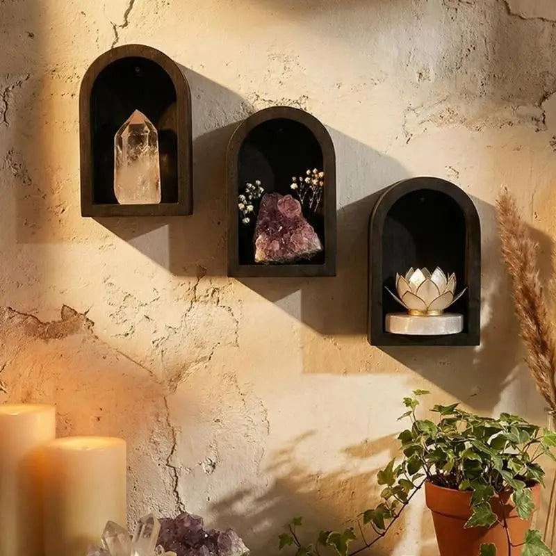 Three dark arched wall shelves mounted on a textured beige wall, styled with a clear crystal, a purple amethyst cluster, and a gold lotus candle holder. Warm candles and a potted plant are visible in the foreground.