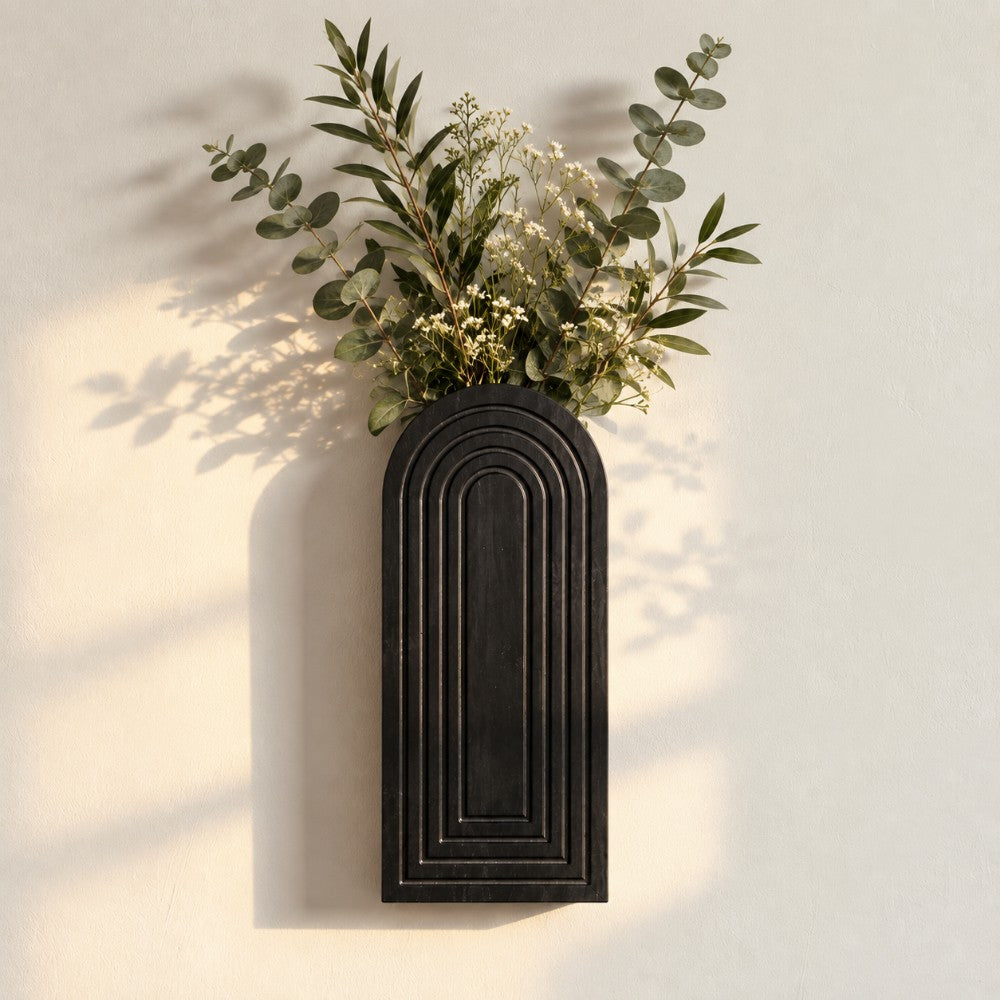 A black arched wall-mounted planter with layered concentric arch detailing, filled with green eucalyptus leaves and small white flowers, mounted on a warm off-white wall with soft leaf shadows.