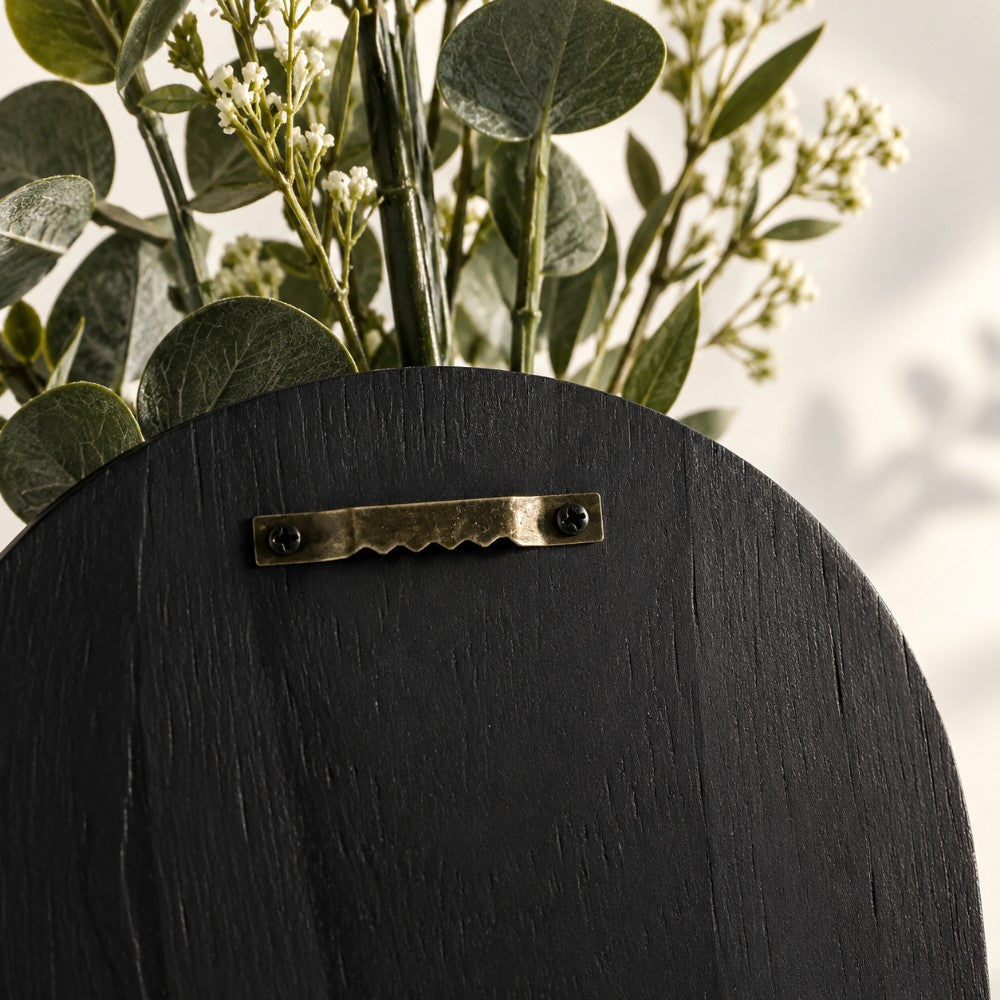 Close-up of a black wooden wall-mounted planter with a brass hook and green eucalyptus stems and white flowers emerging from the top.