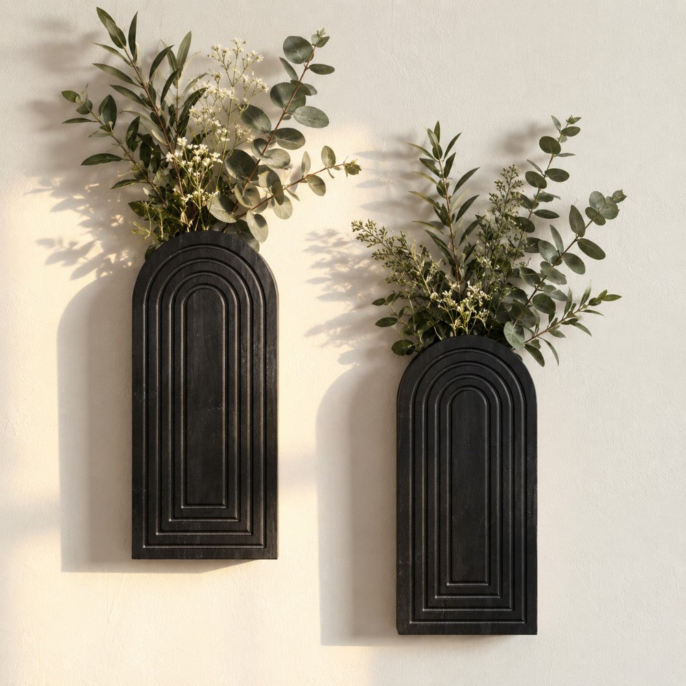 Two black arched wall-mounted planters with ribbed detailing, each filled with green eucalyptus and small white flowers, mounted on a light beige wall.